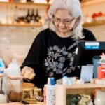 Elderly woman preparing drinks in a cozy coffee shop setting with warm ambiance.